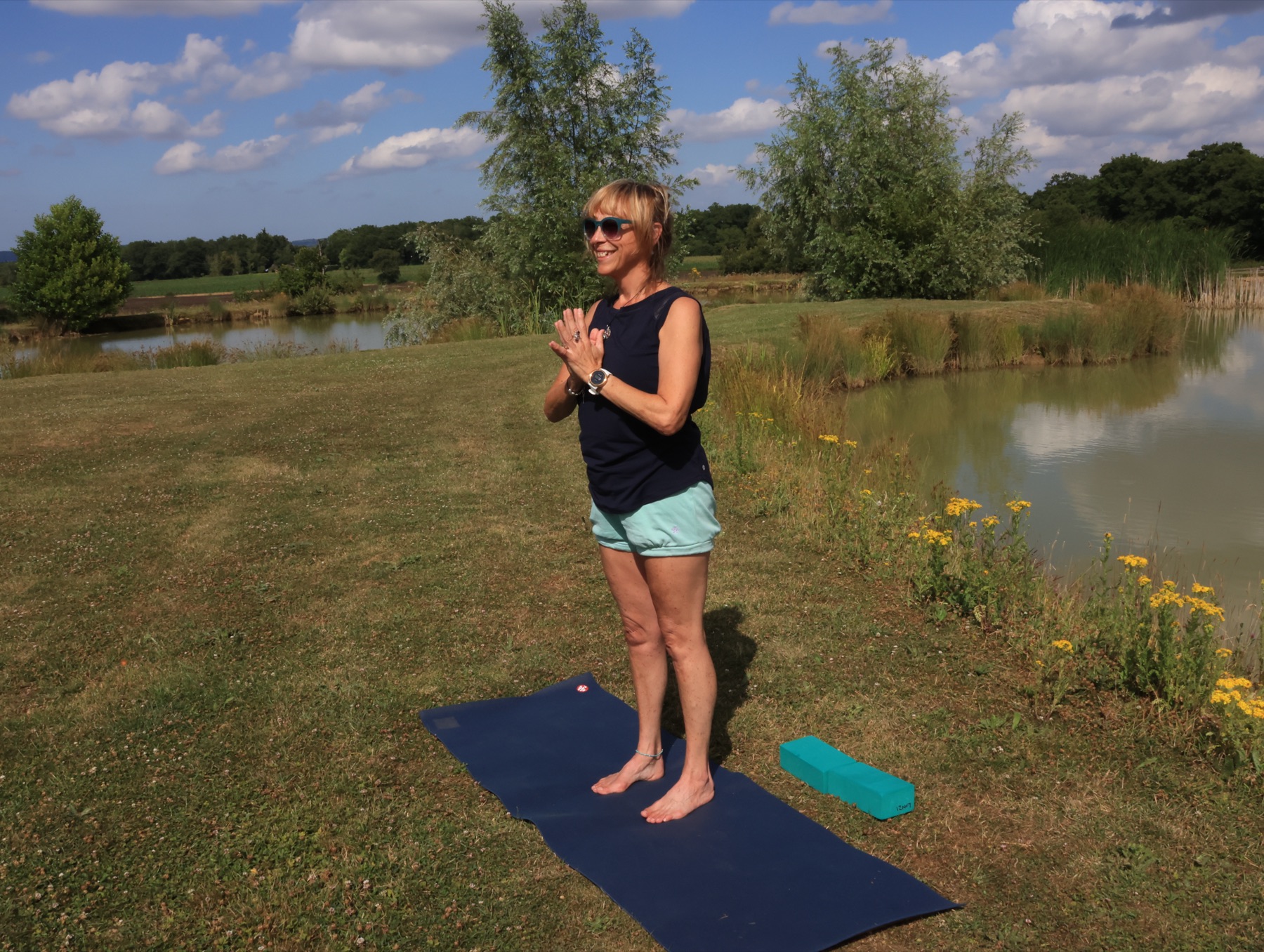 Linzi standing on a navy mat on mown grass beside a lake, hands at heart centre, wildflowers and trees behind