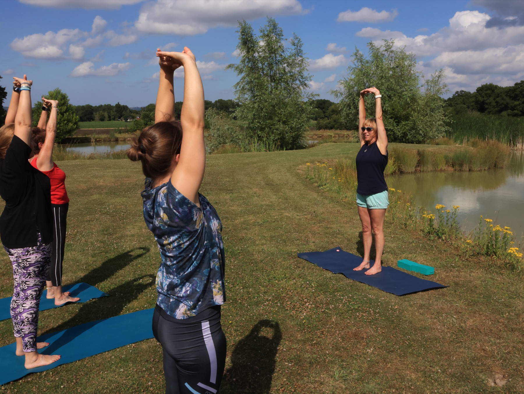Linzi leading an outdoor class: students on blue mats on a lakeside lawn with arms overhead