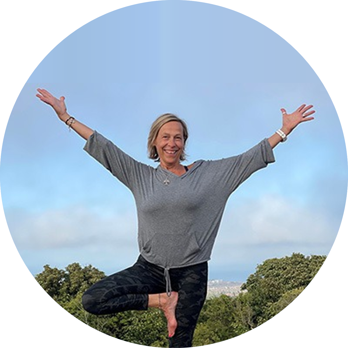 Linzi Kent in tree pose, arms wide, blue sky and treetops behind
